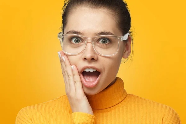 Young female in polo neck sweater and transparent glasses shouting wow with open mouth isolated on yellow background