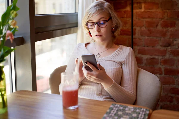 Bella donna guardando lo schermo del telefono, esaminando attentamente le stime delle spese. foto di giovane ragazza con occhiali e vestito beige. Una bella bionda pensa ai prossimi giorni, fa un piano — Foto Stock