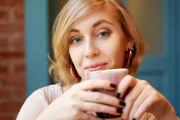 Menina satisfeita com um belo sorriso e cabelo loiro penteado Kare olha para a câmera. Uma jovem mulher bebendo café em um café contra uma parede de tijolo e uma porta azul — Fotografia de Stock