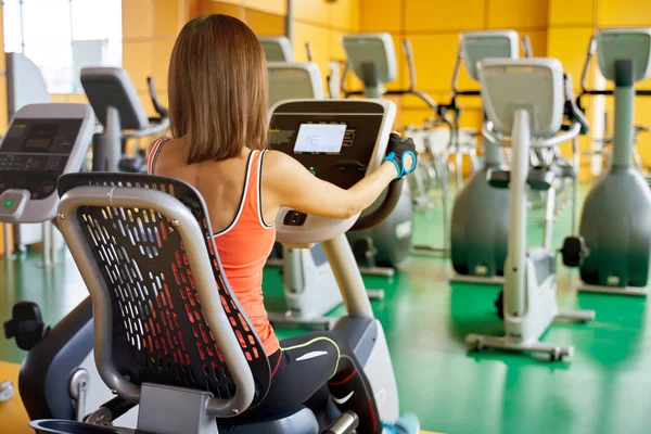 Foto esportiva mulher sentada em uma bicicleta estacionária, fazendo exercício cardio, pedalando, mede o pulso. Silhueta das costas de uma jovem no ginásio — Fotografia de Stock