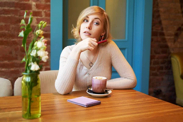 Poważna miło dziewczyna patrząc na kamerę. Młoda kobieta pije kawę w kawiarni z ceglanym murem i niebieskimi drzwiami — Zdjęcie stockowe