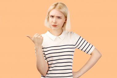 Dejected kız kaşlarını çatma, mutsuz hissediyor, işaret parmağı sola işaret, bazı öğe görmek rahatsız olmak, kopya alanı ile kum bej arka plan üzerinde izole