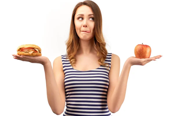 Молодая женщина, держащая яблоко и бургер, на лице вдумчивые и мечтательные эмоции от выбора между здоровой едой и вкусным фаст-фудом. Концепция здорового питания и контроля веса — стоковое фото