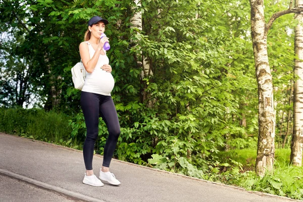 Spor eğitimi sırasında parkta yeşillikler ve ağaçlar arasında eşofman lı ve beyzbol şapkalı hamile bir genç kadın yürüyor.