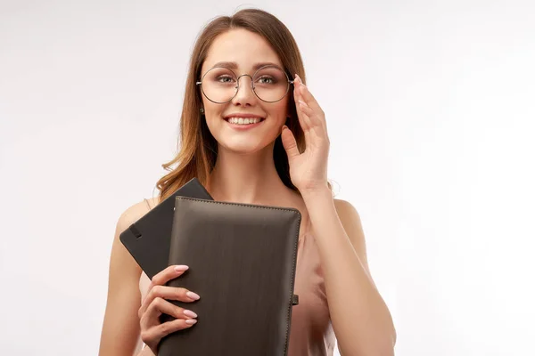 Flicka elev innehar en anteckningsbok och justerar hennes stora runda glasögon och ett vänligt leende brett. Studio närbild porträtt på isolerad vit bakgrund — Stockfoto