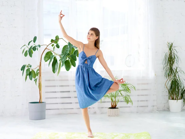 Uma bela mulher loira de pé em uma perna, dedos mostrando Zen. O treinador atraente está usando um vestido azul. Menina praticando manhã alongamento em casa na frente de grande janela — Fotografia de Stock