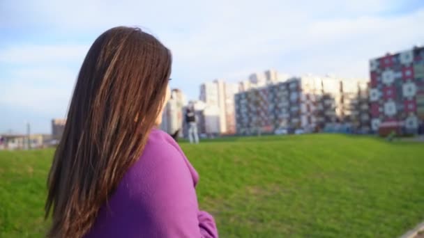 一个阳光灿烂的女人在公园里散步的视频。中年黑发，长发，风中飘扬着紫色格子花，在镜头下转身，你可以看到背像的轮廓 — 图库视频影像
