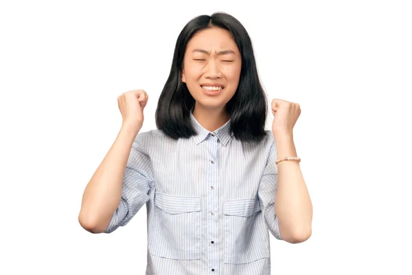 Une jolie femelle serre les poings, lève les mains avec indignation, ferme les yeux, sourit, fronce les sourcils, colère, irritation, stress. Jeune femme vêtue de chemise de bureau rayée se tient isolé contre le mur blanc Image En Vente