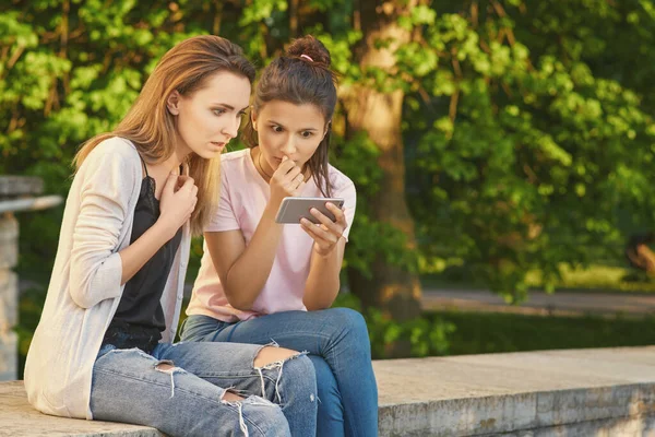 A barátnők telefonálgatnak, vakációznak, felidézik a szép pillanatokat. Gyönyörű nők hétköznapi ruhákban a parkban ülve. A nők meglepődnek a hírek sokkolta tartalom és a tájékoztatás Jogdíjmentes Stock Fotók