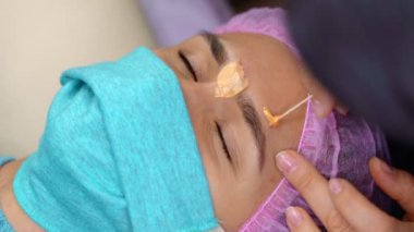 Tıbbi maskeli kadın kaşlarını alıyor. Güzel bir kadının kaş düzeltme prosedürü uygulanır. Güzellik salonunda kanepede yatan kız. Yakın çekim müşteri yüzü ve kozmetik uzmanı elleri. 4K video
