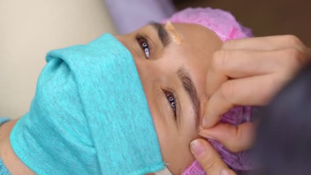 Γυναίκα με ιατρική μάσκα που γυαλίζει τα φρύδια. Όμορφη γυναίκα παίρνει φρύδια διαδικασία διόρθωσης. Κορίτσι ξαπλωμένο στον καναπέ στο σαλόνι ομορφιάς. close-up πελάτη πρόσωπο και εξειδικευμένη cosmetologist χέρια. Βίντεο 4K — Αρχείο Βίντεο