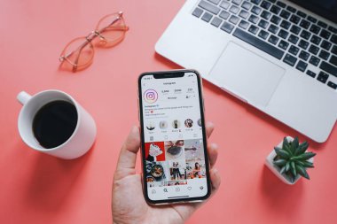 El tutarak Apple iPhone XS ile dizüstü bilgisayara karşı ekranda Instagram uygulaması ile pembe arka planda dizüstü bilgisayar ve kahve bardağı 