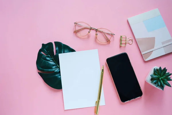 workspace desk with minimal stationery and smartphone with paper and tropical leaf on pink color background