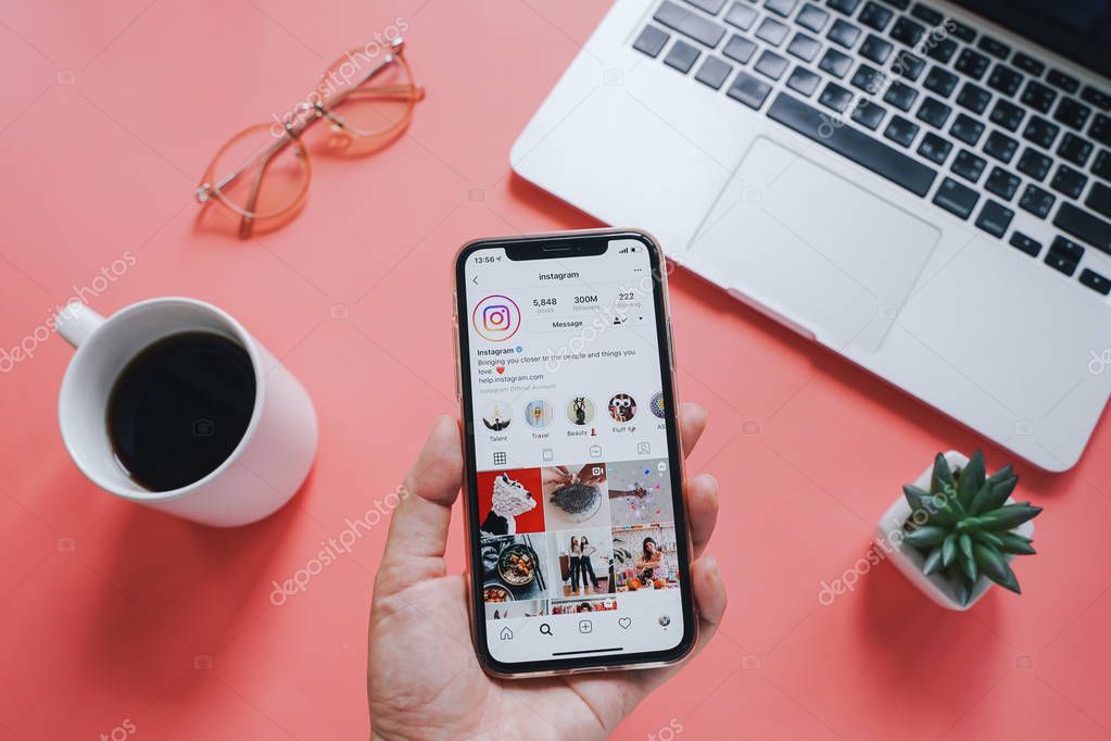 Hand holding Apple iPhone XS with Instagram application on screen against laptop with laptop and coffee cup on pink background