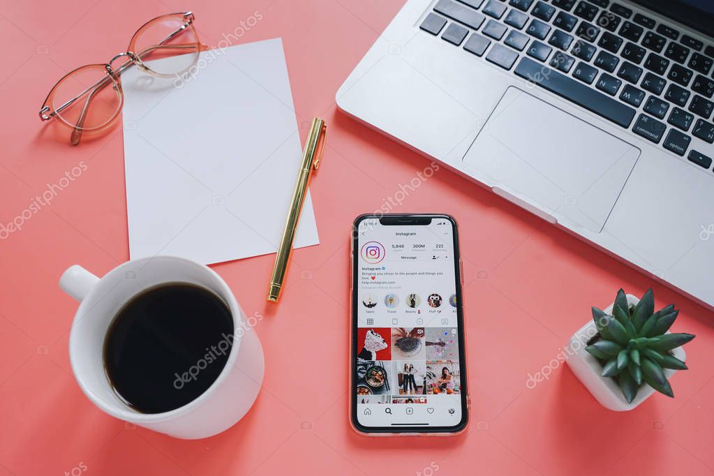 Flat lay of workspace desk and Apple iPhone XS with Instagram application on screen against laptop with laptop and coffee cup on pink background