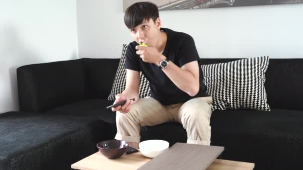 Hombre Joven Sentado Sofá Usando Control Remoto Mientras Come Manzana — Vídeo de stock