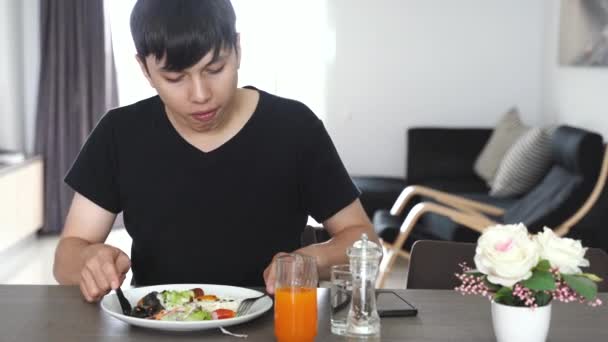 若いアジアの混合人種の男が健康的な朝食を食べ 健康的なライフスタイルでアパート 食べ物や飲み物でオレンジジュースを飲む — ストック動画