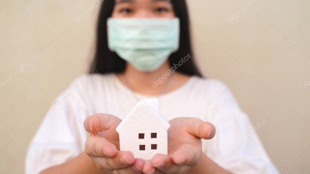 Young asian millennial gen z woman holding white house model and wearing mask for self quarantine or stay at home concept during COVID-19 and coronavirus pandemic