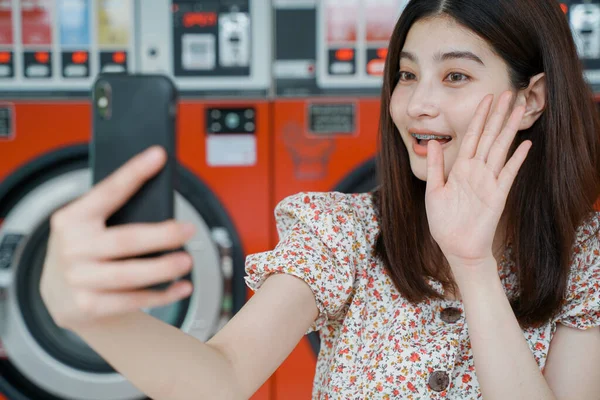 Young asian millennial woman video call with her friend from smartphone during waiting clothes washing at laundromat, lifestyle and technology concept