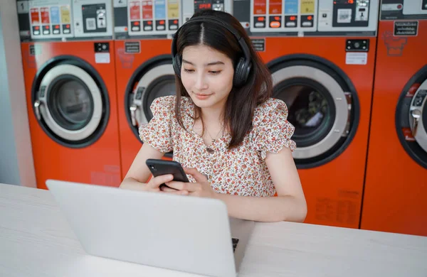 Young asian millennial woman using laptop and listening to music during waiting clothes at laundromat shop, lifestyle and working remotely concept