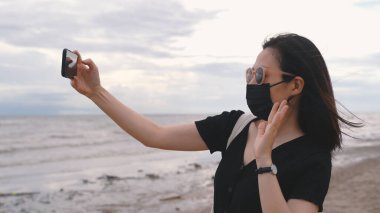 Tıbbi maske takan genç Asyalı bir kadın veya Coronavirus 'tan sonra sahilde akıllı telefon ile görüntülü görüşme, yeni normal yaşam tarzı ve seyahat balonu konsepti