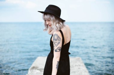 Güzel genç kadın dövme ve deniz arka plan üzerinde poz siyah elbise ile