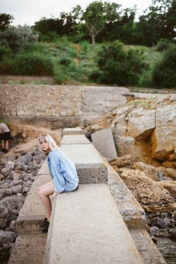 Taşlı sahilde denize sıfır kot ceket giyen güzel genç sarışın hippi kız. Açık havada
