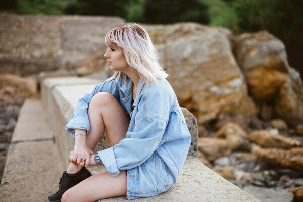Красивая молодая блондинка хипстерша в джинсовой куртке на каменистом пляже у моря. На открытом воздухе
