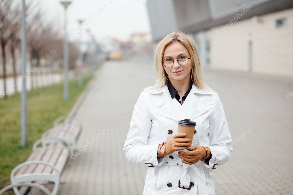 Portrait of successful business woman holding cup of hot drink in hand on her way to work on city street. Beautiful woman with coffee cup near office building.