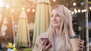 Şehir sokakta kahve ve cep telefonu fincan ile muhteşem genç kadın. Modern akıllı telefon cihazını kullanarak şık mutlu hipster kız.