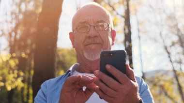 Aktif yaşlı adam parkta yürüyüş ve akıllı telefon kullanarak.