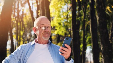 Aktif yaşlı adam parkta yürüyüş ve akıllı telefon kullanarak.