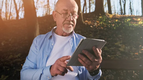 Active senior man walking in the park and using a tablet sitting in the park on the bench.