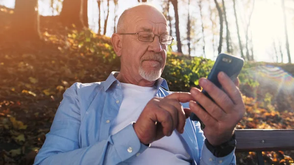 Active senior man walking in the park and using smartphone sitting in the park on the bench.