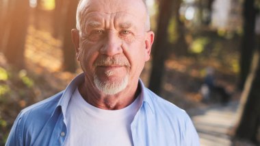 Parkta yürüyen aktif kıdemli adam. Güneş ışığı altında dışarıda dururken gülümseyen sakallı yaşlı adamın portresi.