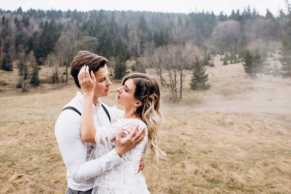 Retrato de la joven pareja caucásica atractiva boda de pie en el prado brumoso en las colinas del campo y abrazándose mientras se miran con amor . — Foto de Stock