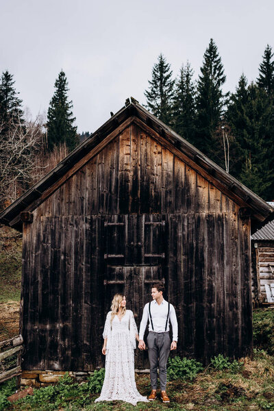 Portrait of the young Caucasian good looking, stylish and happy fiance and fiancee in the wedding clothes standing together, holding hands and looking at each other at the rural wooden building.