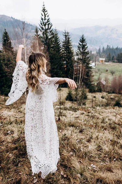 Vista posterior de la joven novia caucásica en el vestido largo blanco de pie en el hermoso claro en las montañas rurales en el nublado día de otoño. Parte trasera . — Foto de Stock