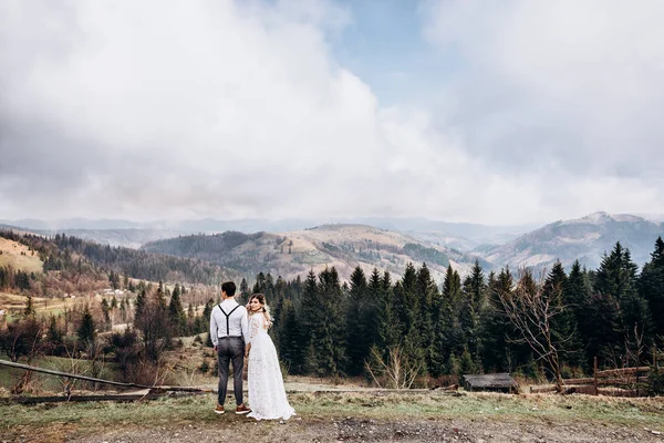 Traseiro do casal de casamento em pé no lugar muito pitoresco nas montanhas. A noiva a olhar para a câmara por cima do ombro. Visão traseira . — Fotografia de Stock