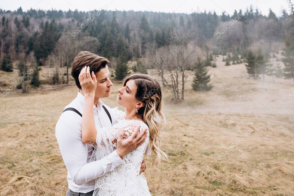 Portrait shot of the young Caucasian attractive wedding couple standing at the misty meadow in the countryside hills and hugging while looking at each other with love.