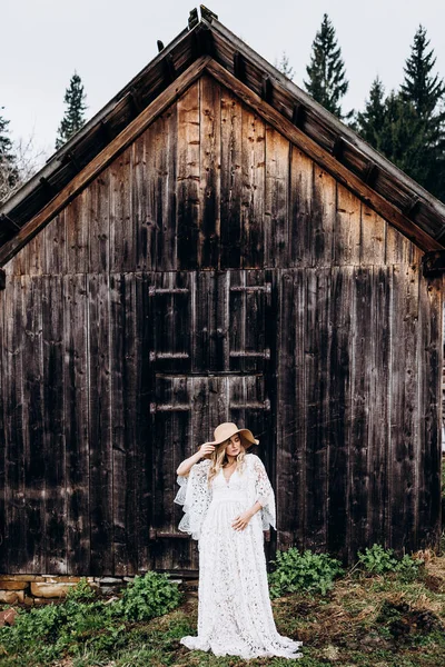 Portrait de la jeune mariée blonde caucasienne dans la longue robe blanche et grand chapeau debout et posant à la place rurale à l'ancien bâtiment en bois. Extérieur . — Photo