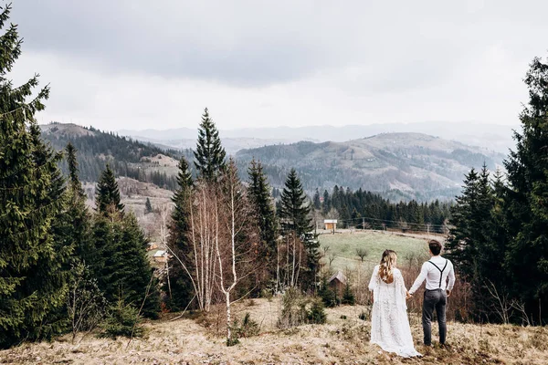 Tillbaka syn på den kaukasiska romantiska paret på deras bröllopsdag under fotograferingen i de lantliga bergen, hålla händerna och titta på den pittoreska naturen. Bakre. — Stockfoto