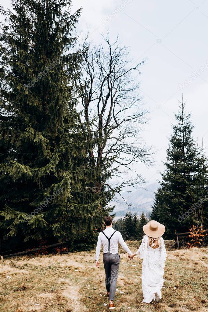 Back view on the Caucasian young just married couple walking hand in hand the countryside meadow with pines. Woman in the long while dress and big hat. Rear. Outdoor.