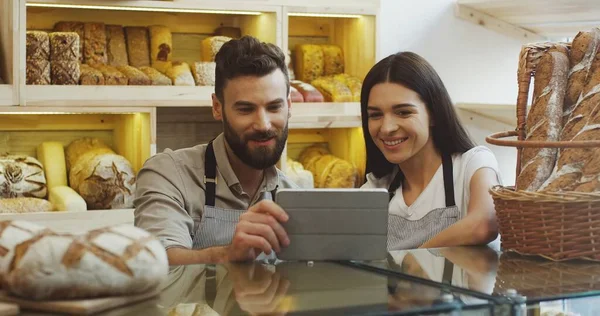 Attractive male and female bread vendors scrolling and watching something on the tablet device while standing at the counter in the bakery shop.