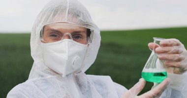 Portrait of Caucasian beautiful young female biologist researcher in protective costume, mask and goggles holding flask with chemicals and smiling to camera in field.