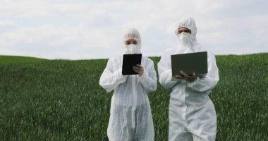 Koruyucu kostümlü beyaz erkek ve kadın bilim adamları yeşil alanda ellerinde tablet, dizüstü bilgisayar ve konuşmalarla dikiliyorlar. Erkek ve kadın biyologlar hasatla uğraşıyorlar..