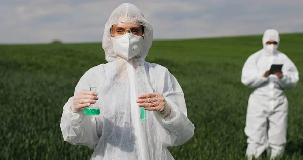 코카서스 여성 과학자가 살충제 액체가 담긴 보호 복을 입고 현장에서 실험을 하고 있습니다. 화학 물질을 가지고 일하는 실험실의 남녀 일꾼들. — 스톡 사진