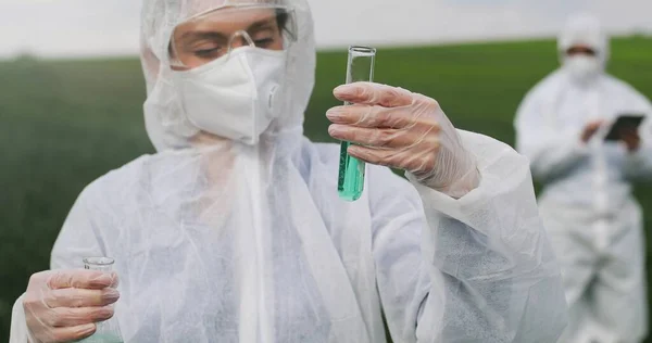 Kaukasische Wissenschaftlerin in Schutzkleidung, die Schläuche mit flüssigen Pestiziden hält und auf dem Feld experimentiert. Laborarbeiterinnen und -arbeiter arbeiten mit chemischer Substanz. — Stockfoto