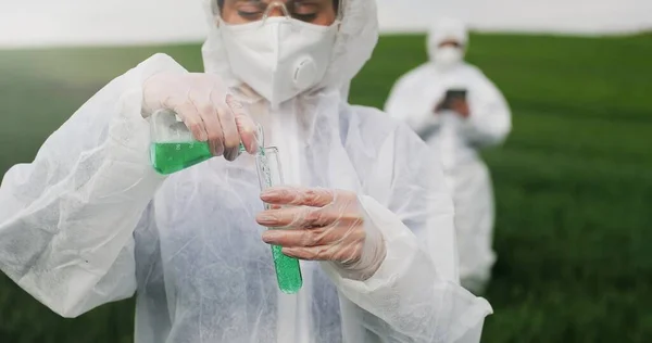 Закріпіть трубку з хімічною рідиною в руках жіночого кавказького дослідника, який стоїть на зеленому полі. Використання сільськогосподарських пестицидів. Жінка в захисному костюмі контролює боротьбу зі шкідниками.. — стокове фото