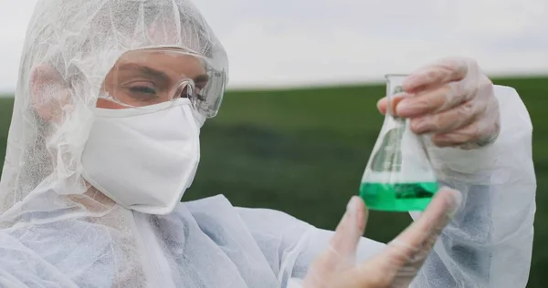 Retrato de branco bela jovem bióloga pesquisadora em traje protetor, máscara e óculos segurando frasco com produtos químicos e sorrindo para a câmera no campo . — Fotografia de Stock